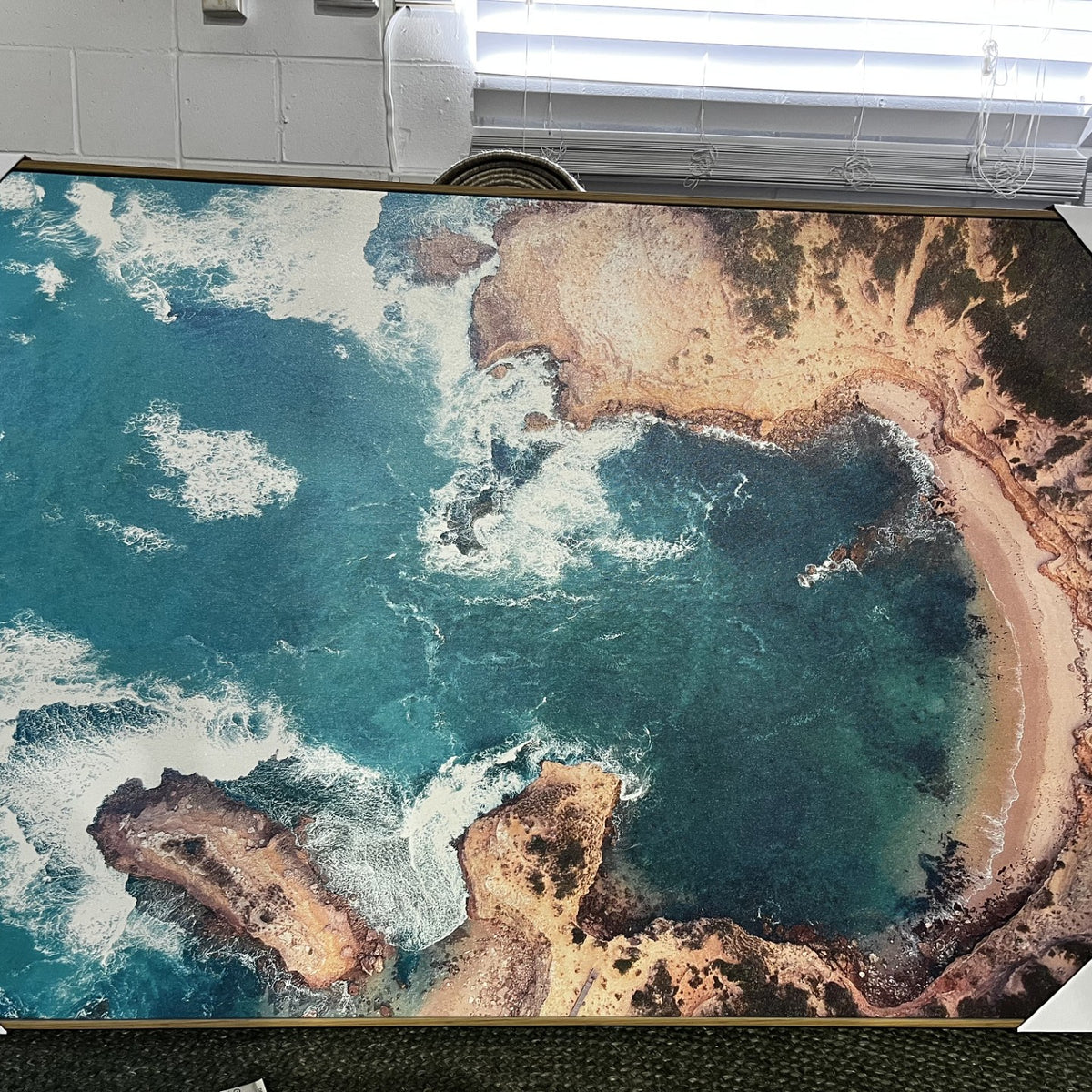 A large framed aerial photograph of a coastal bay with turquoise water, white surf, and sandy shoreline, displayed indoors with corner protectors on a gallery-wrapped canvas.