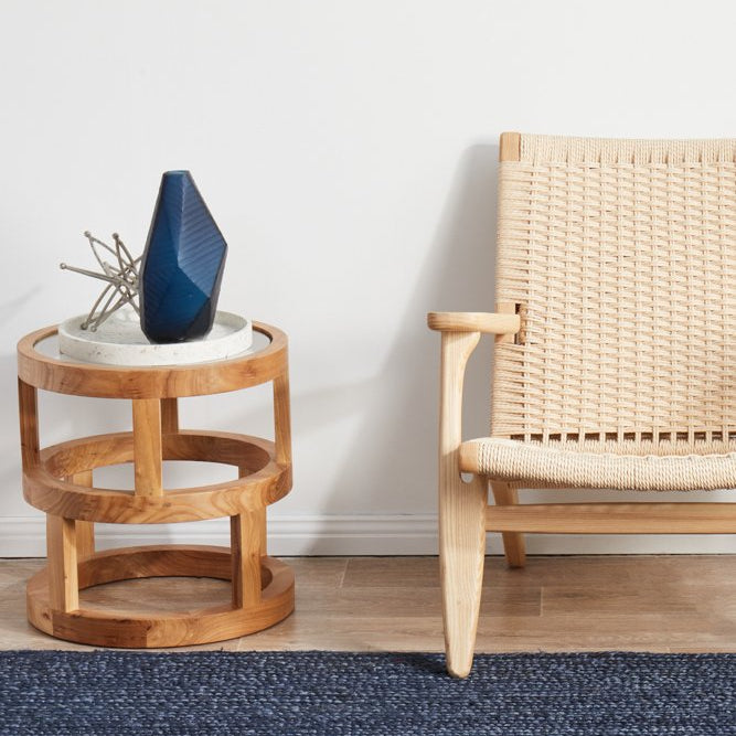 Wooden side table with a blue vase and plant, next to a woven chair on a blue rug.