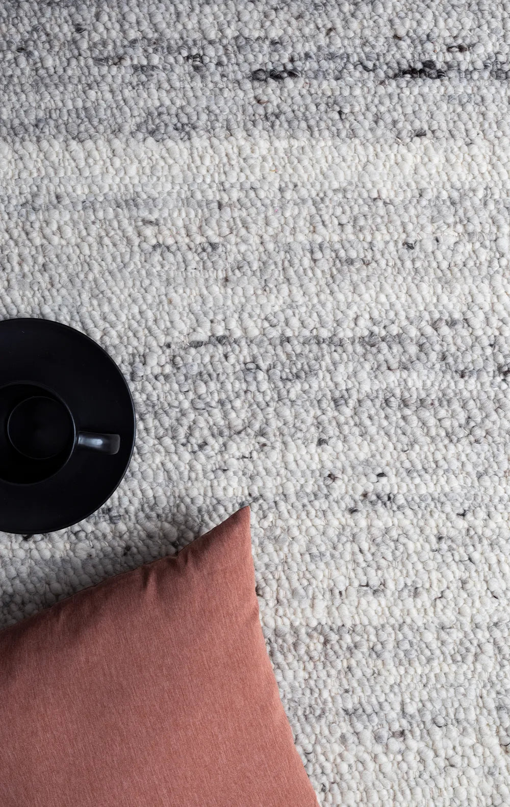 Close-up of a textured gray rug with a black object and pink pillow.