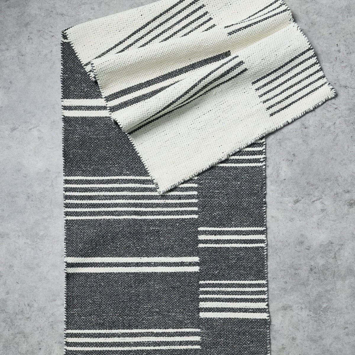 A folded woven textile with a black and off-white striped geometric pattern, displayed on a textured gray concrete surface.