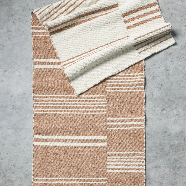 A woven rectangular throw blanket with a striped pattern in warm tan and cream, folded diagonally on a textured gray concrete surface.
