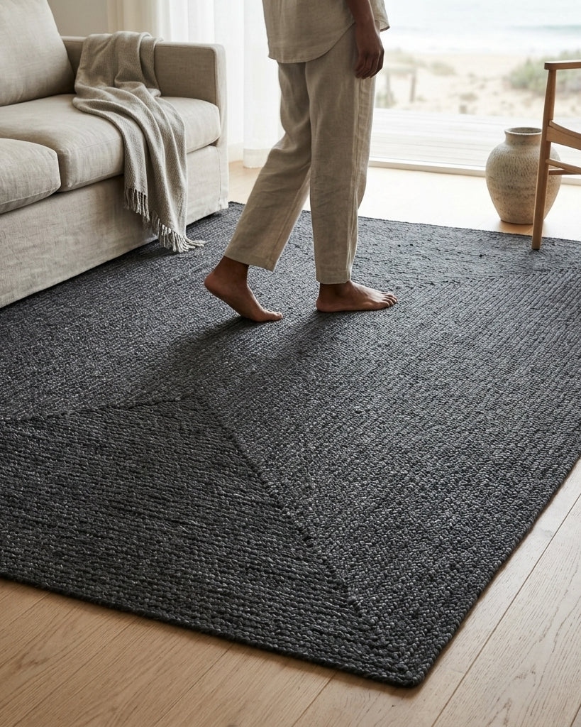 A dark charcoal gray textured area rug on a light wood floor in a bright living room, with a person standing barefoot on the rug near a beige sofa and chair.