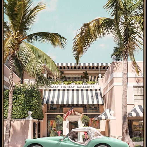 A framed art print depicting a retro mint green convertible car parked on a street in front of a building with a black and white striped awning, framed by tall palm trees under a light blue sky.
