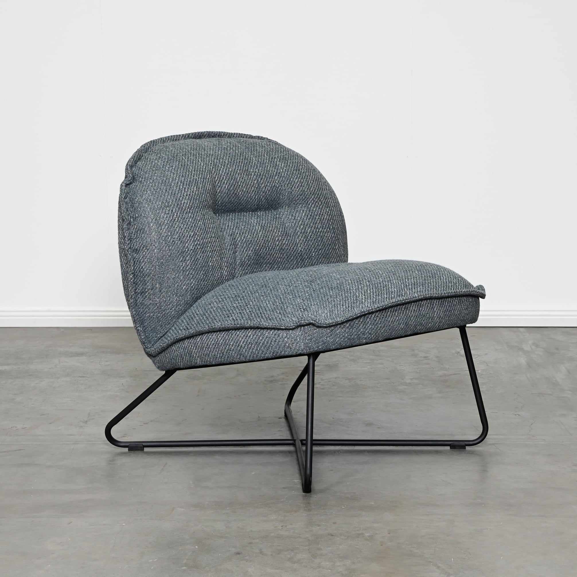 A modern low-profile accent chair upholstered in a textured gray fabric with a black metal frame, set against a minimalist white wall and gray concrete floor.