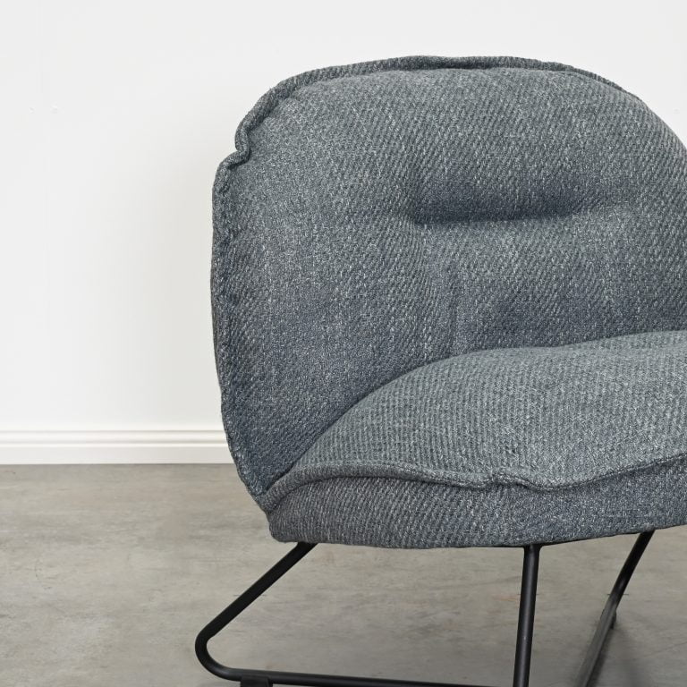 A close-up of a modern upholstered accent chair with a textured gray fabric and a black metal wire frame, set against a plain white wall and concrete floor.