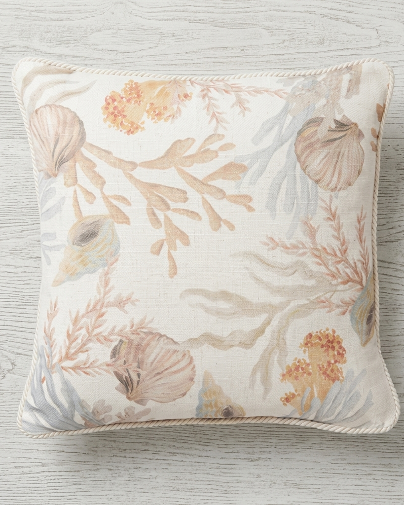 A square decorative throw pillow with a nautical design featuring seashells, coral, and seaweed in muted beige, peach, and light blue-gray tones on an off-white background, resting on a light gray wood plank surface.