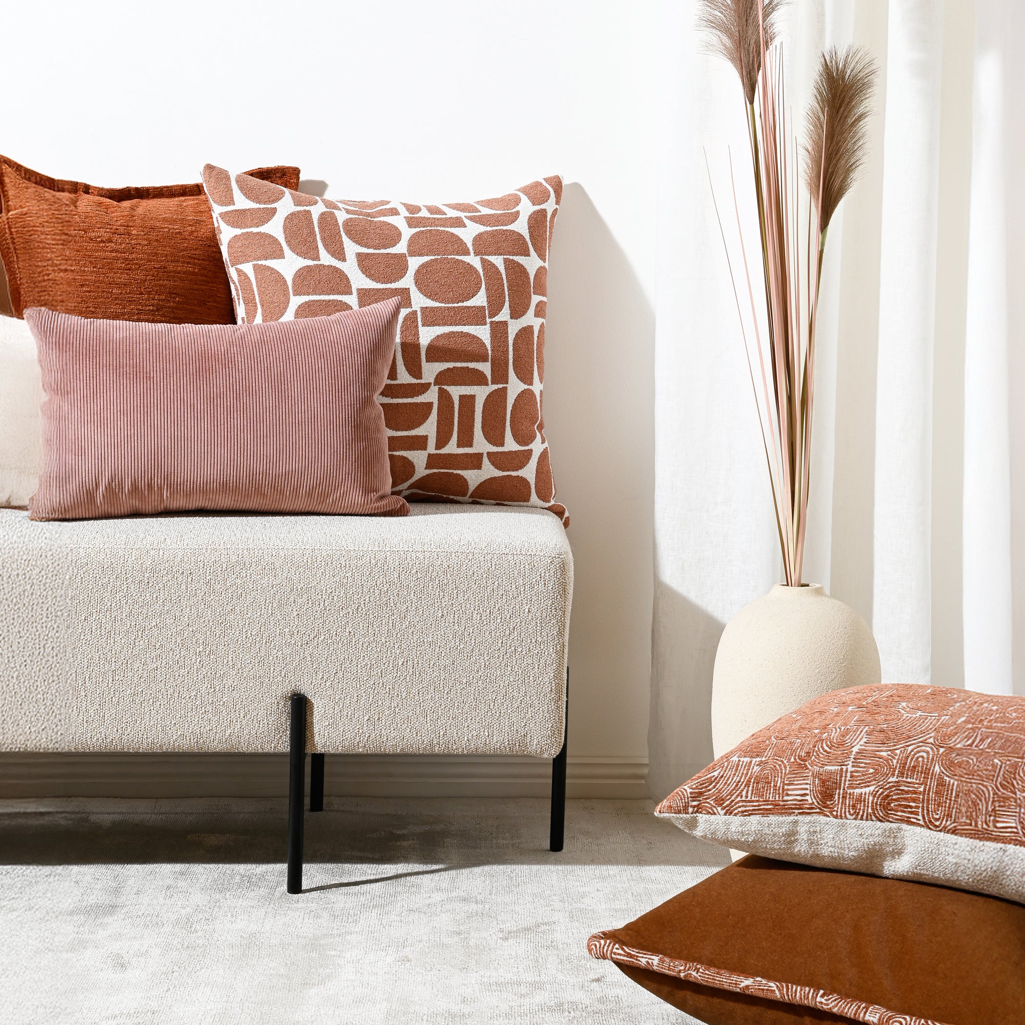 A styled interior scene with a light upholstered bench or sofa holding several decorative throw pillows in rust, dusty pink, and cream-and-brown patterned fabric. A tall white vase with dried grass stands against a white wall, and part of another cushion is visible in the foreground on a light rug.