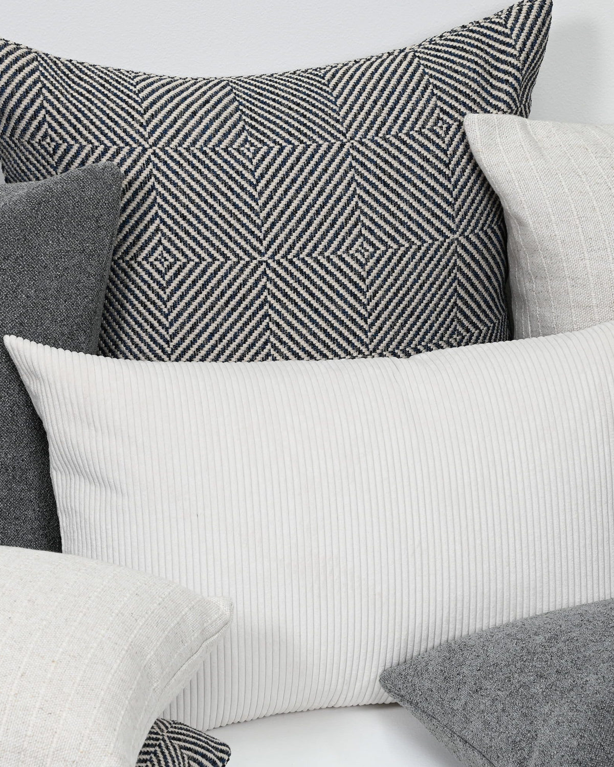 A styled arrangement of multiple decorative throw pillows in neutral tones, including a prominent white ribbed pillow in the foreground, a black-and-white geometric patterned pillow behind it, and several gray and off-white pillows.