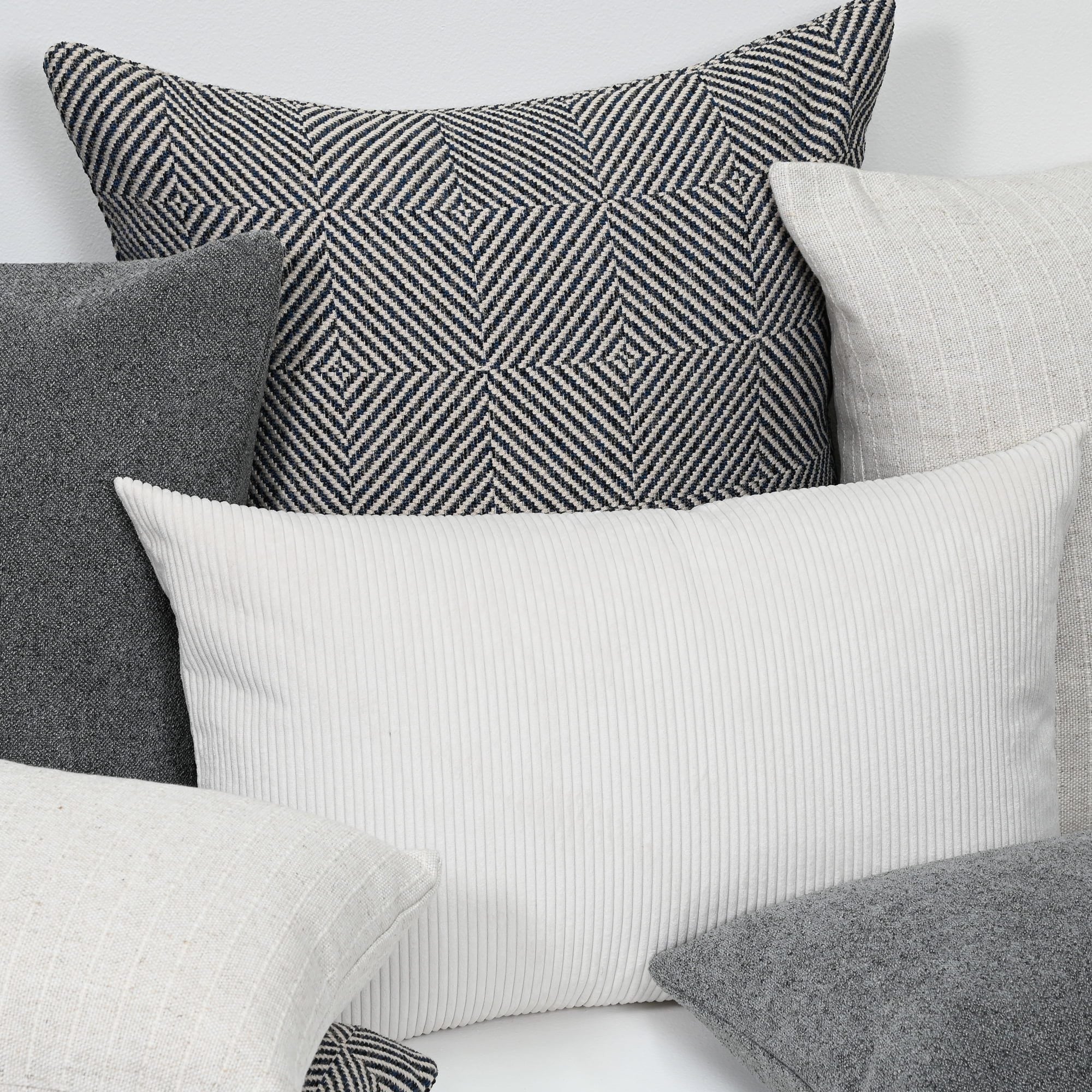 A styled arrangement of multiple decorative throw pillows in neutral tones, including a prominent white ribbed pillow in the foreground, a black-and-white geometric patterned pillow behind it, and several gray and off-white pillows.