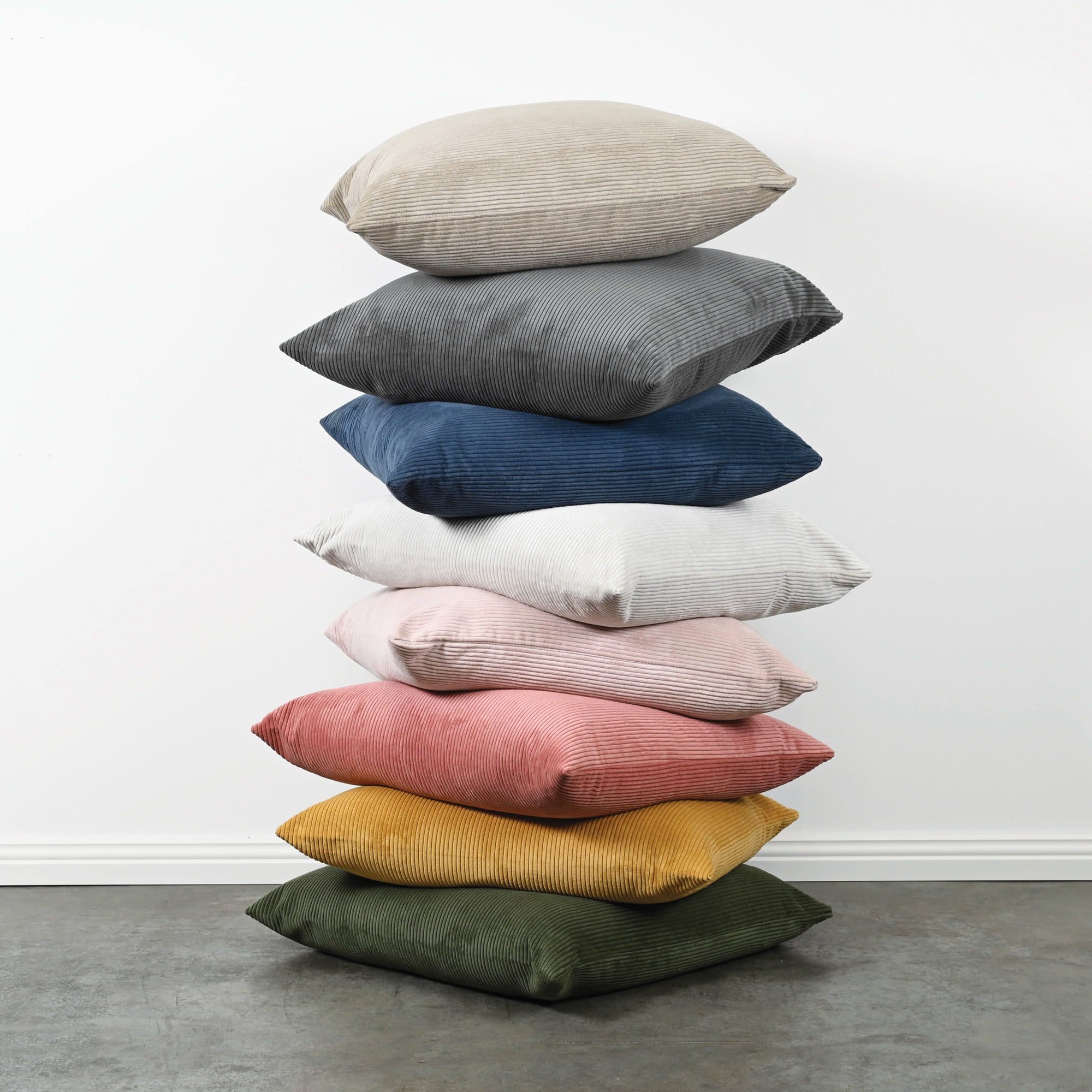 A stack of nine throw pillows in various colors, including beige, gray, navy, white, light pink, dusty pink, mustard yellow, and olive green, arranged on a gray floor against a white wall.