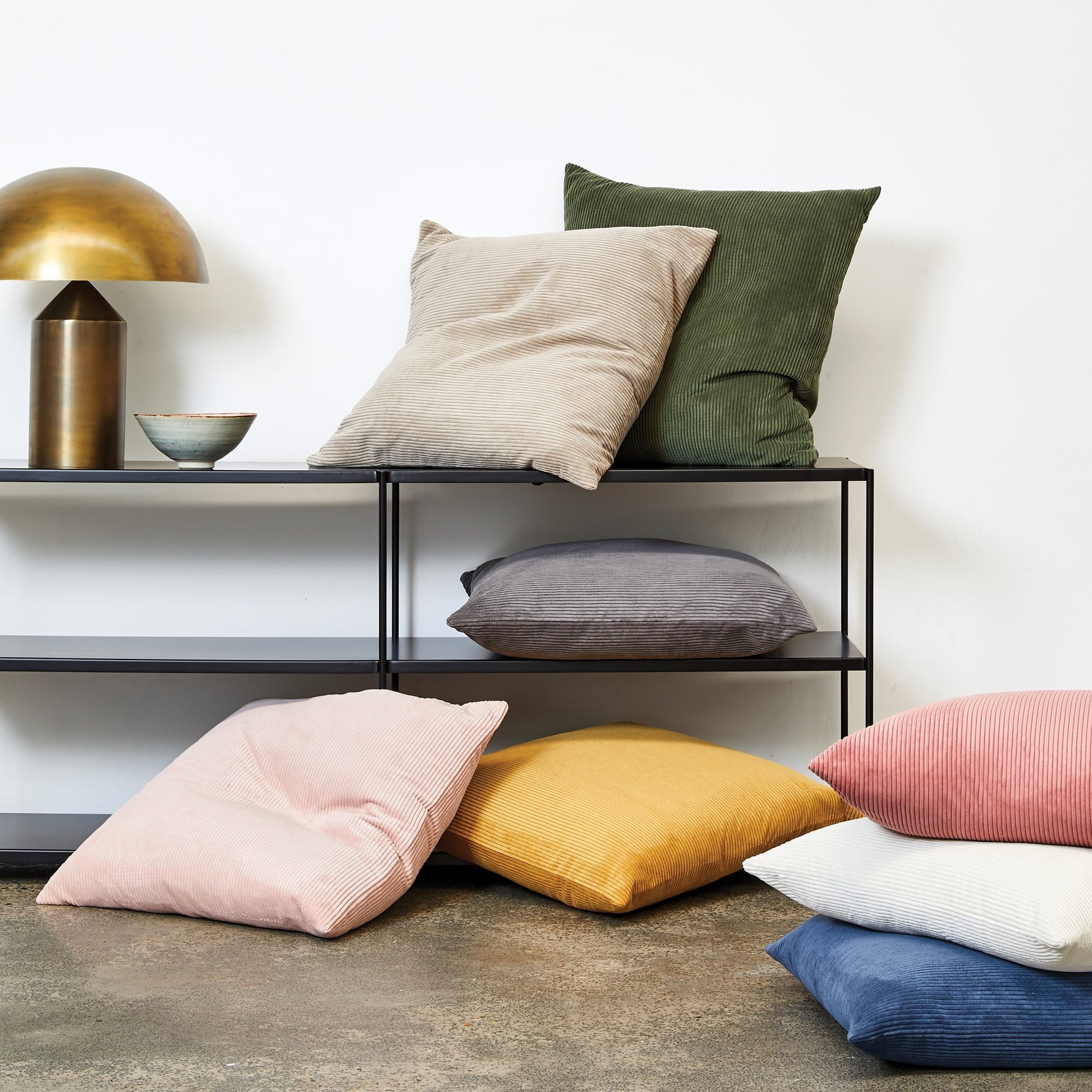 A collection of textured throw pillows in various colors including beige, olive green, gray, pink, yellow, white, and blue, styled on a black metal shelf and the floor against a white wall.