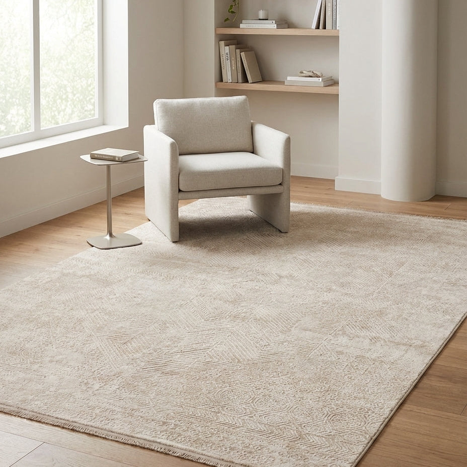 A large light beige area rug with a subtle textured pattern, shown in a bright, modern room with a white armchair, a small side table, and wooden flooring.