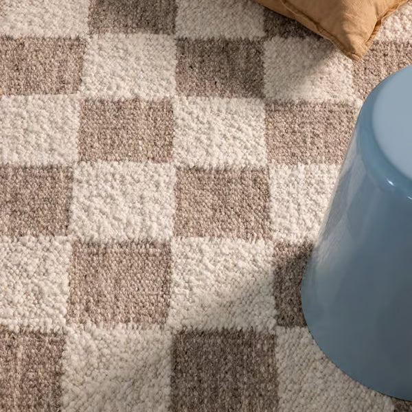A close-up of a textured area rug with a beige, cream, and brown checkerboard pattern, styled with a tan pillow and a light blue stool.