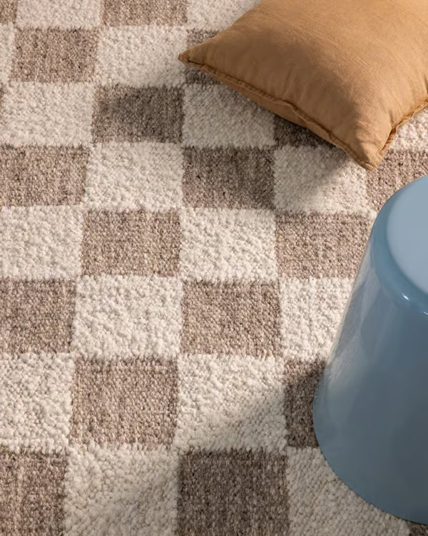 A close-up of a textured area rug with a beige, cream, and brown checkerboard pattern, styled with a tan pillow and a light blue stool.