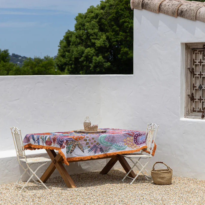 Outdoor setting with a table and chairs under a colorful tablecloth, against a white wall with a window.