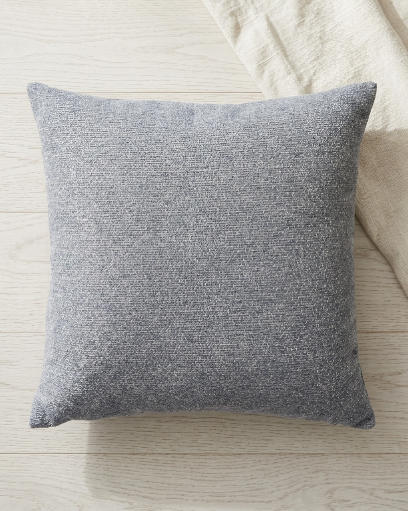 A square gray textured throw pillow resting on a light wood floor next to a cream linen fabric.