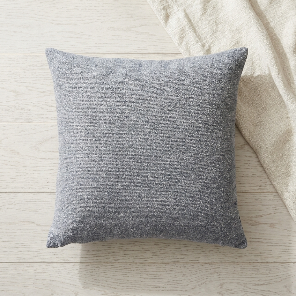 A square gray textured throw pillow resting on a light wood floor next to a cream linen fabric.