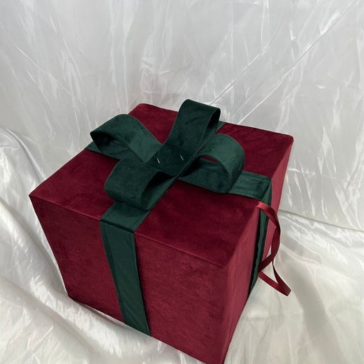 A square gift box wrapped in deep red velvet fabric and tied with a dark green satin ribbon and bow, set against a wrinkled white backdrop.
