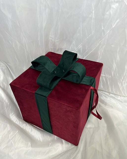 A square gift box wrapped in deep red velvet fabric and tied with a dark green satin ribbon and bow, set against a wrinkled white backdrop.