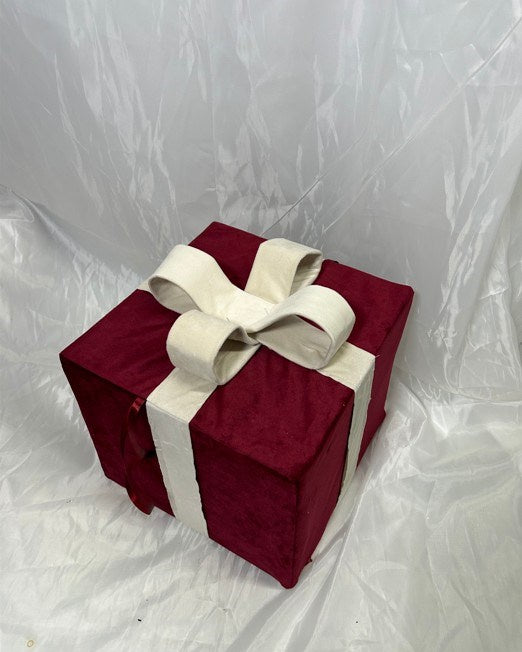 A square gift box wrapped in deep burgundy velvet fabric with a wide cream ribbon tied into a large bow on top, photographed against a wrinkled white backdrop.