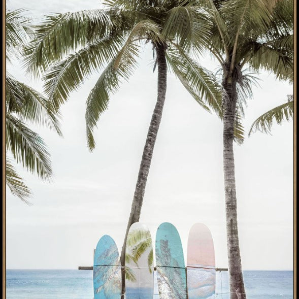A framed art print depicting a tropical beach scene with three palm trees, ocean waves, and three colorful surfboards resting on a wooden bench in the foreground.
