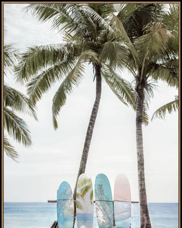 A framed art print depicting a tropical beach scene with three palm trees, ocean waves, and three colorful surfboards resting on a wooden bench in the foreground.