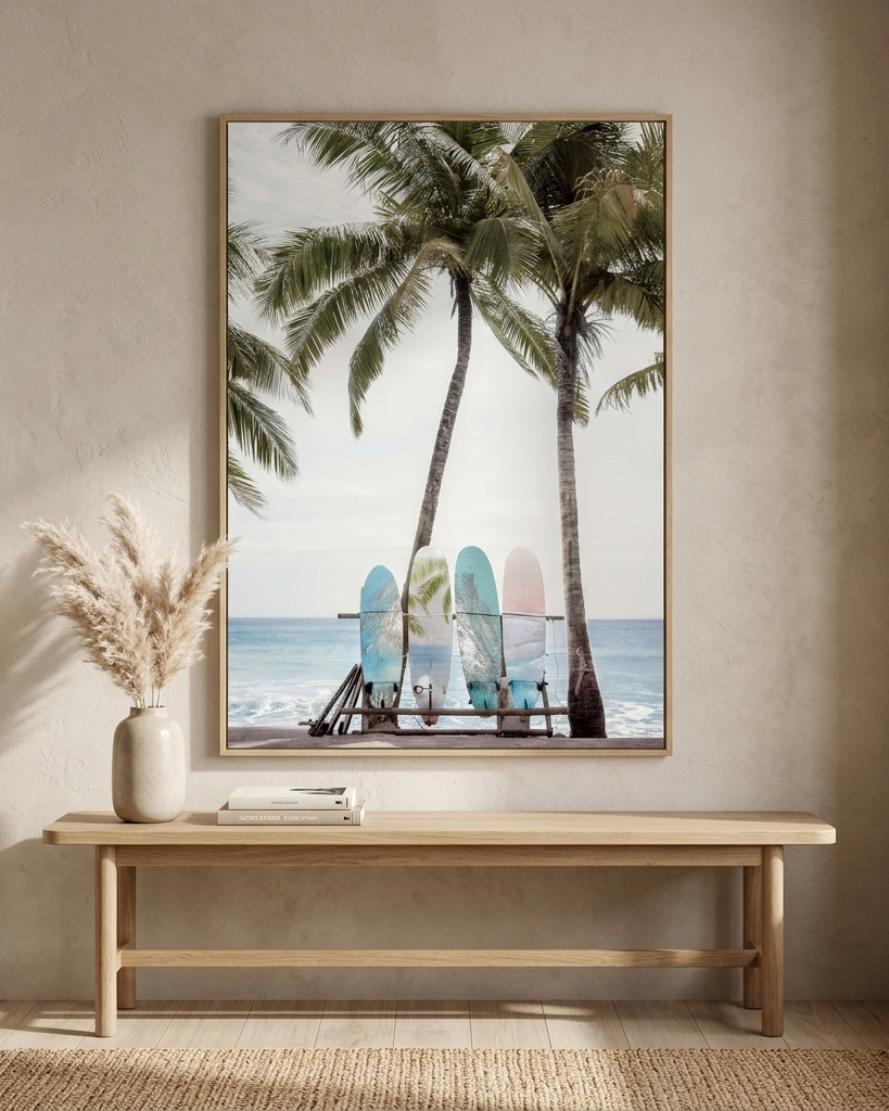 A framed vertical art print depicting a tropical beach scene with palm trees and colorful surfboards, displayed on a beige wall above a light wood bench with a vase of dried grass.