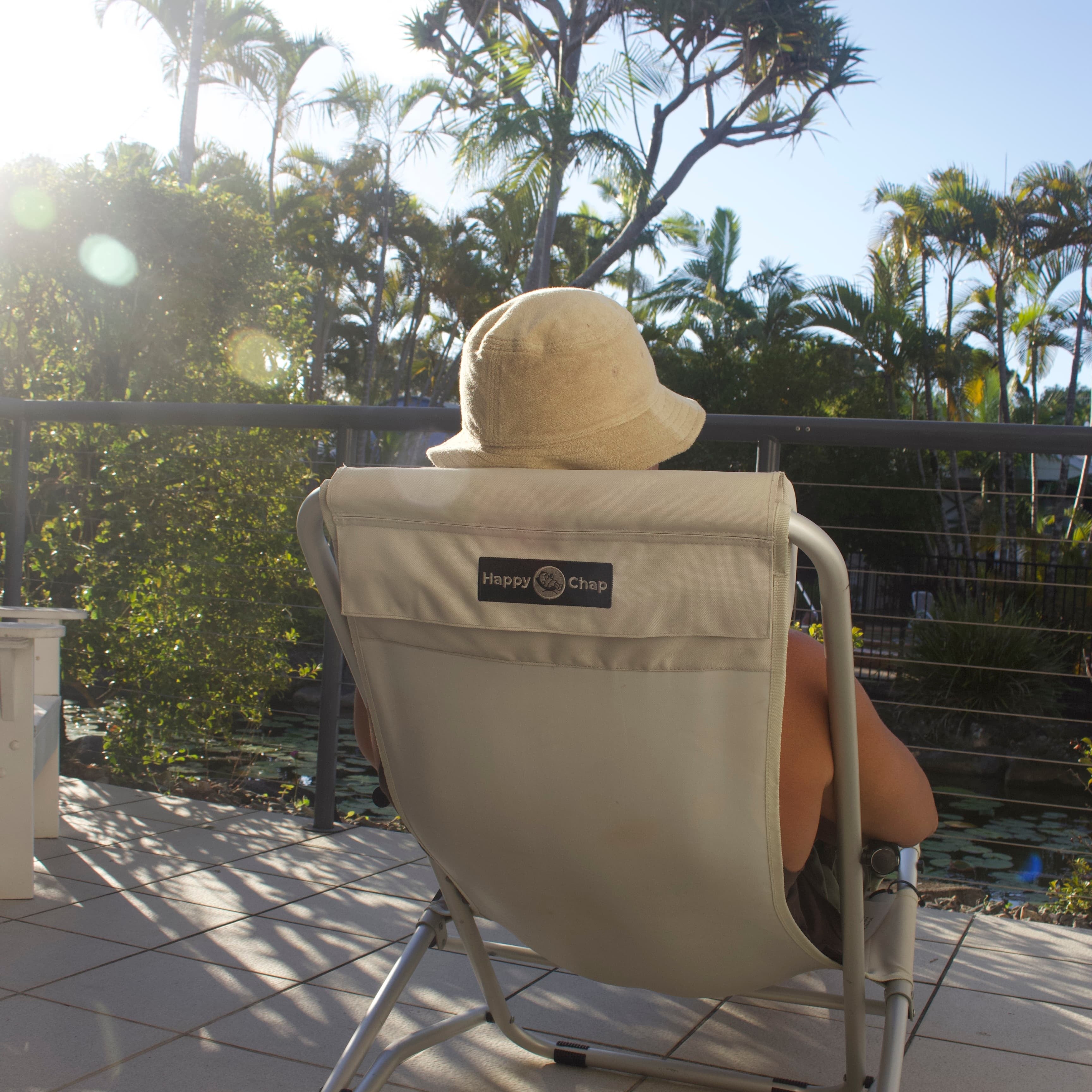 Person sitting in a beige chair with a logo, wearing a wide-brimmed hat, on a tiled patio with palm trees in the background.