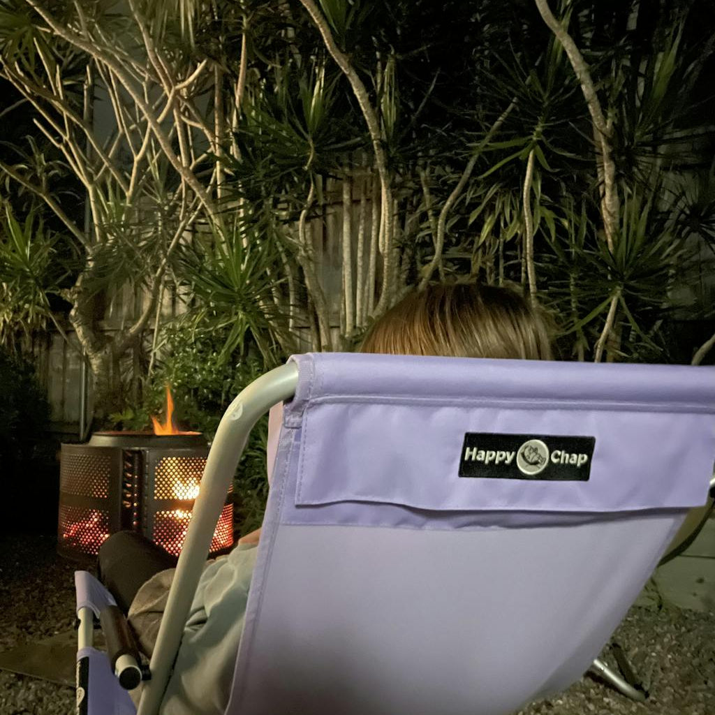 Person sitting in a purple chair with 'Happy Chap' logo near a fire pit at night.