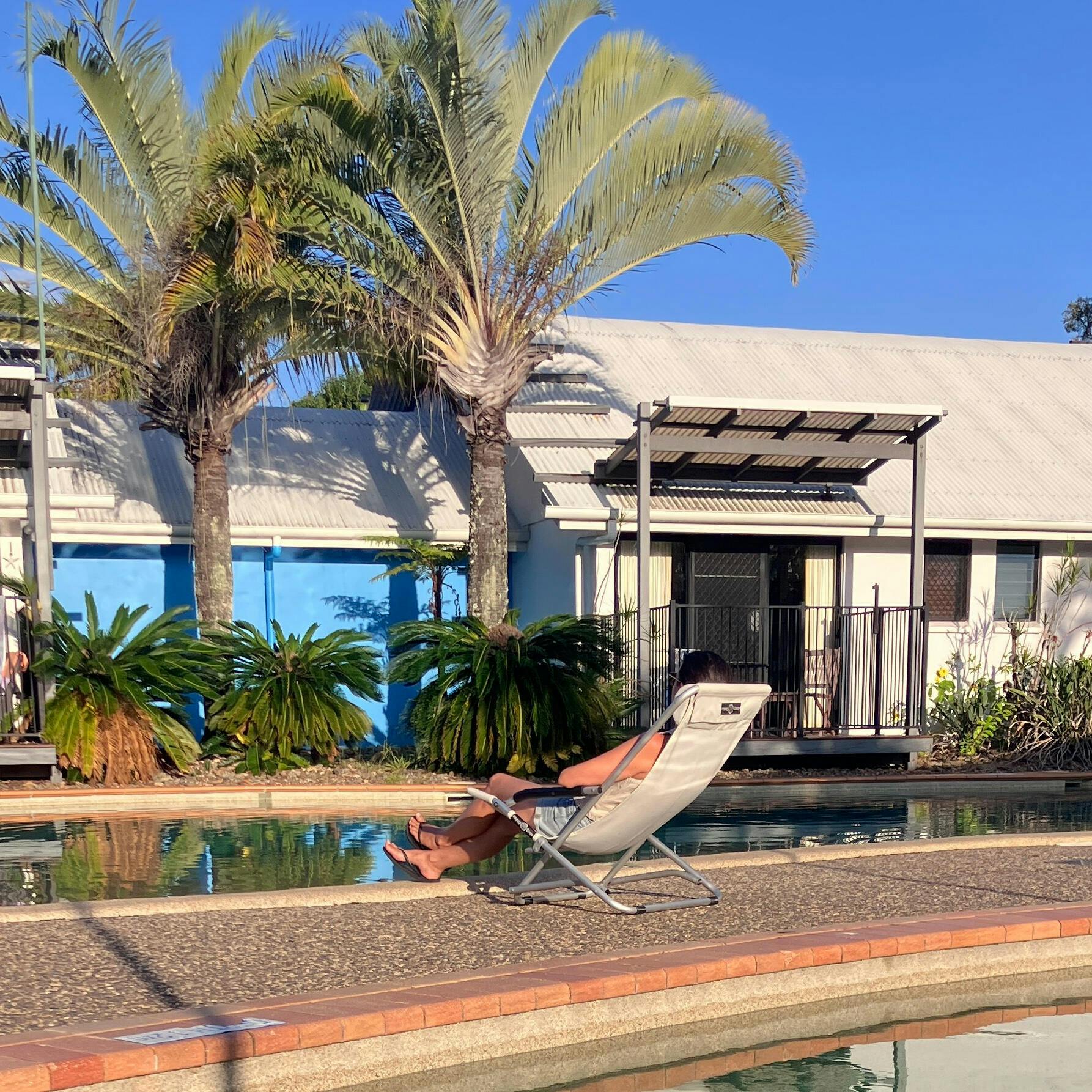 Person lounging by a pool with palm trees and a house in the background