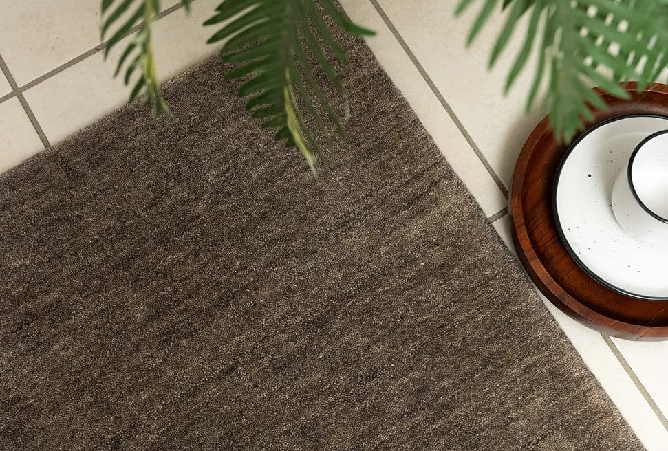 A rectangular emerald grey wool rug placed on a tiled floor with a round wooden object and green foliage in the background.