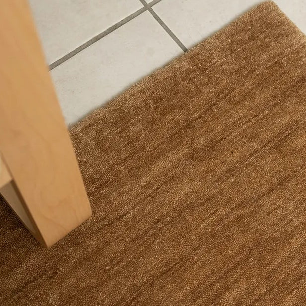 Close-up view of a honey brown wool rug with a textured, chunky woven style, placed on a tiled floor next to a wooden furniture leg.