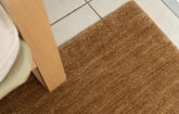 Close-up view of a honey brown wool rug with a textured, chunky woven style, placed on a tiled floor next to a wooden furniture leg.