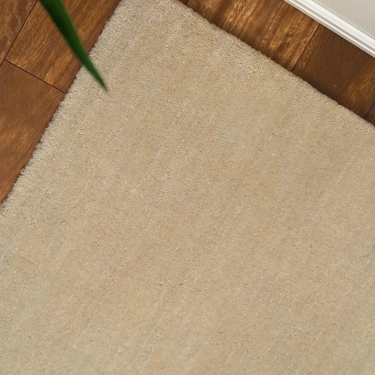 A close-up image of a beige wool rug with a textured weave pattern, placed on a wooden floor with a plant partially visible to the side.