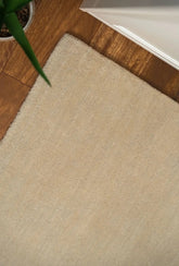 A close-up image of a beige wool rug with a textured weave pattern, placed on a wooden floor with a plant partially visible to the side.