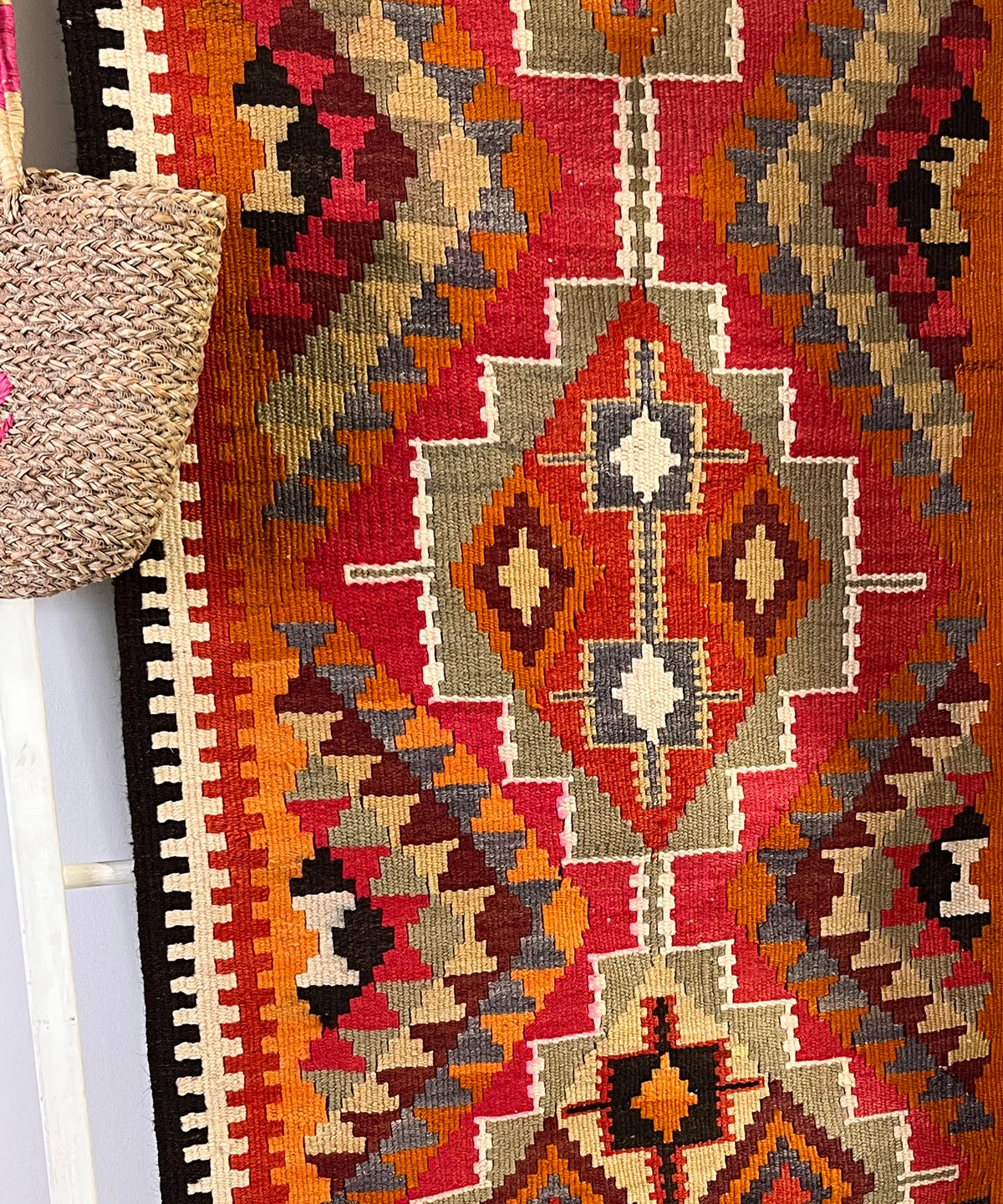 A close-up of a woven geometric runner rug with a Southwestern or Navajo-inspired pattern in red, orange, beige, brown, black, and gray, draped over a white ladder with a woven straw bag hanging on the left.