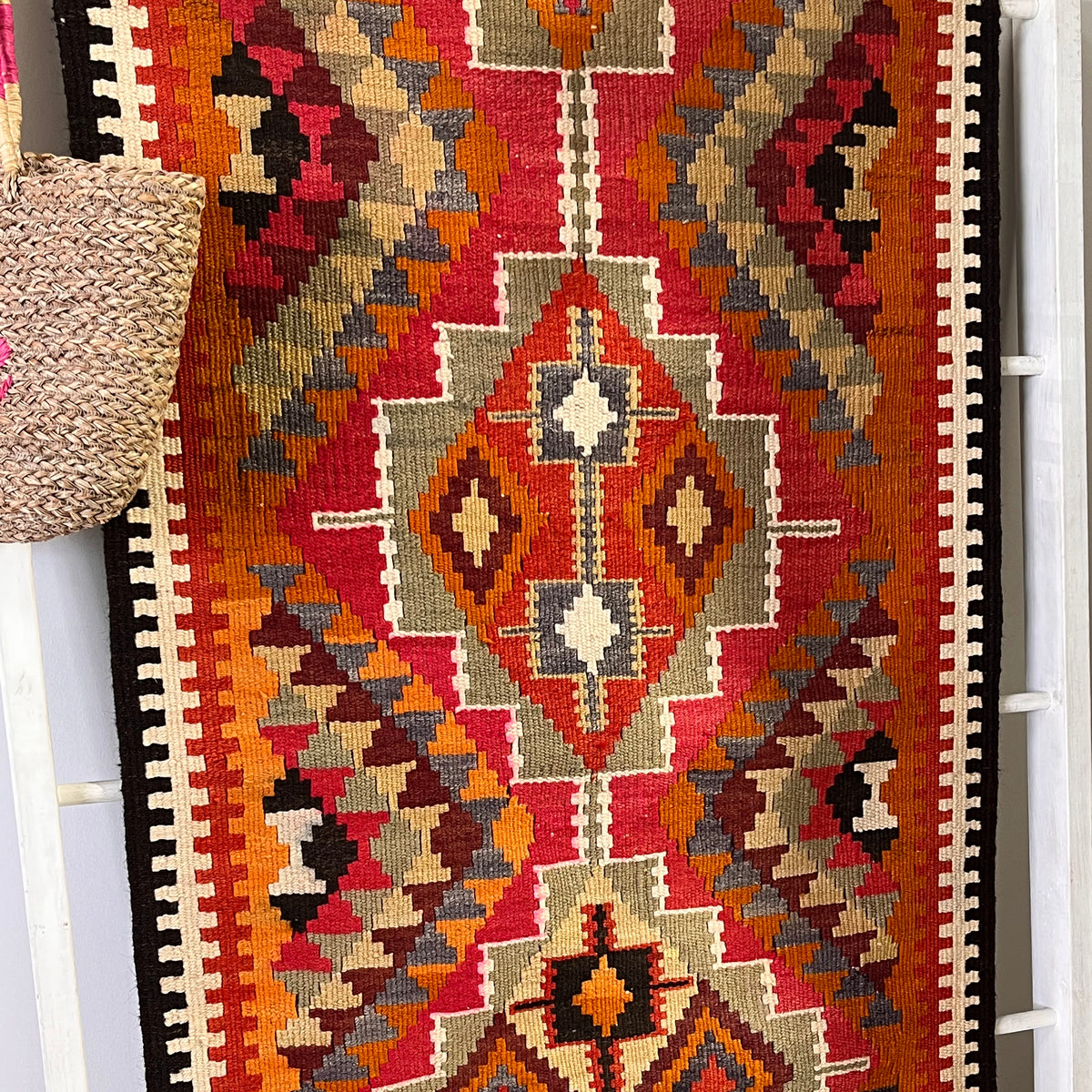 A close-up of a woven geometric runner rug with a Southwestern or Navajo-inspired pattern in red, orange, beige, brown, black, and gray, draped over a white ladder with a woven straw bag hanging on the left.