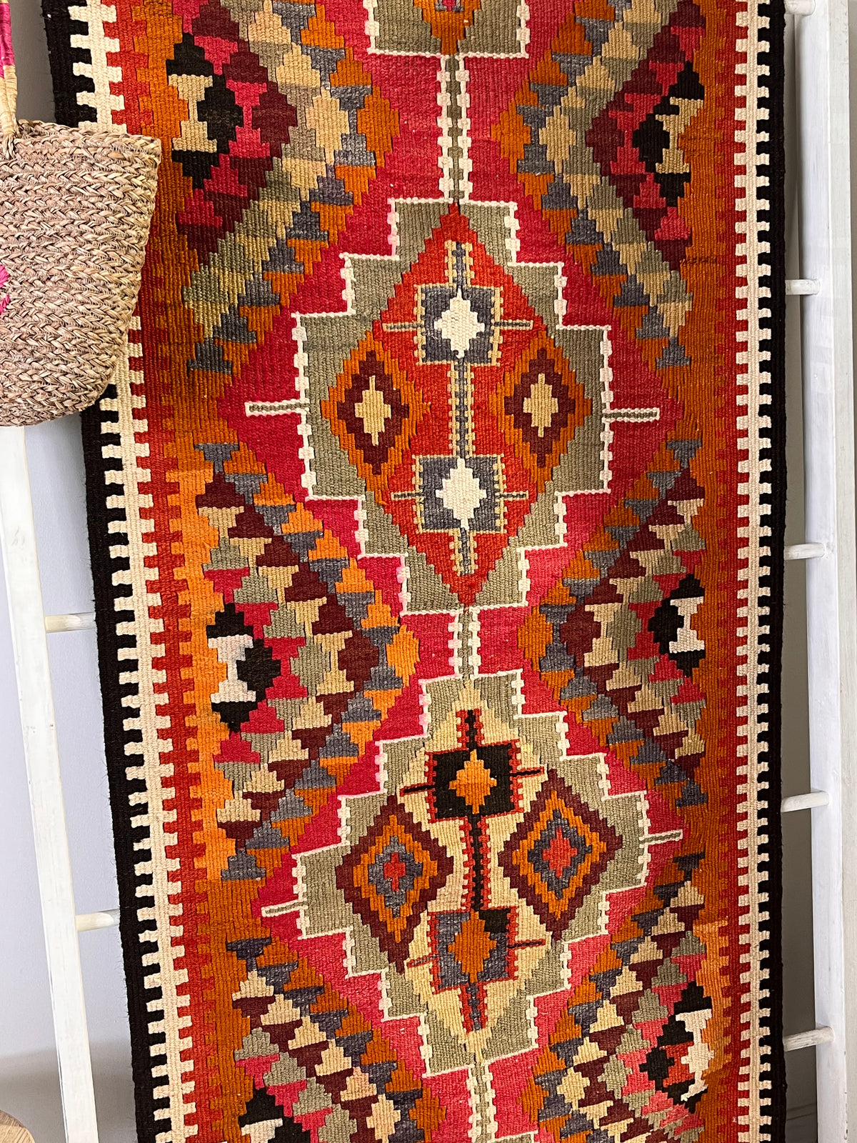 A close-up of a woven geometric runner rug with a Southwestern or Navajo-inspired pattern in red, orange, beige, brown, black, and gray, draped over a white ladder with a woven straw bag hanging on the left.