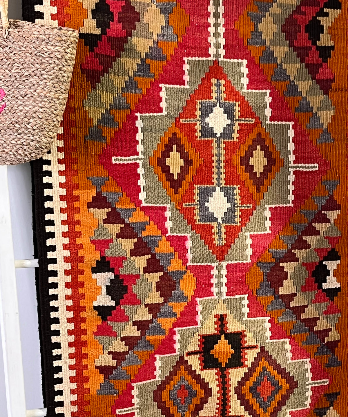 A vintage-style kilim runner rug with a geometric tribal pattern in red, orange, brown, beige, and black, hanging on a white rack.