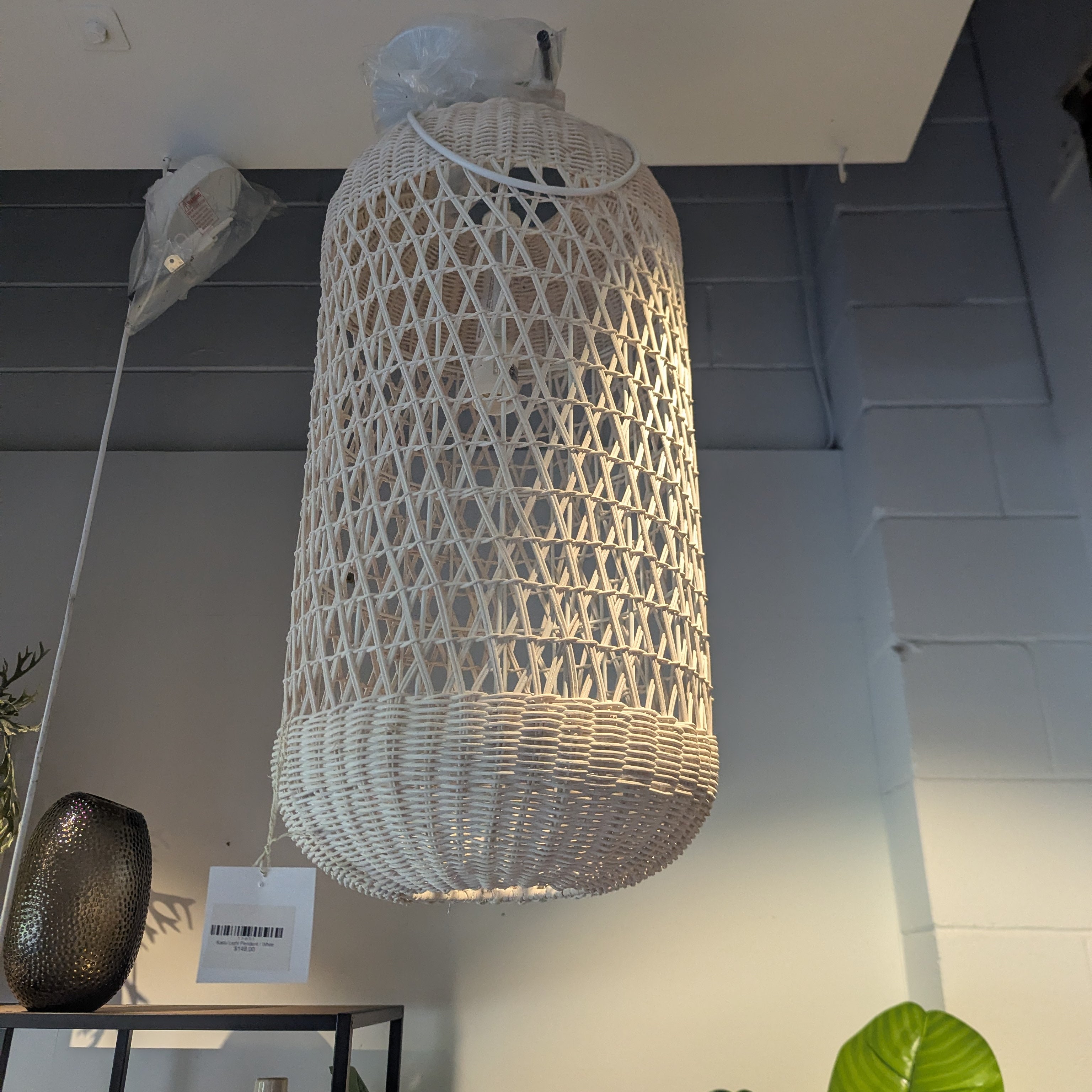 A large cylindrical pendant light made of woven natural fiber, hanging from a ceiling in an indoor showroom. The lamp has an open lattice top and a tightly woven lower shade.