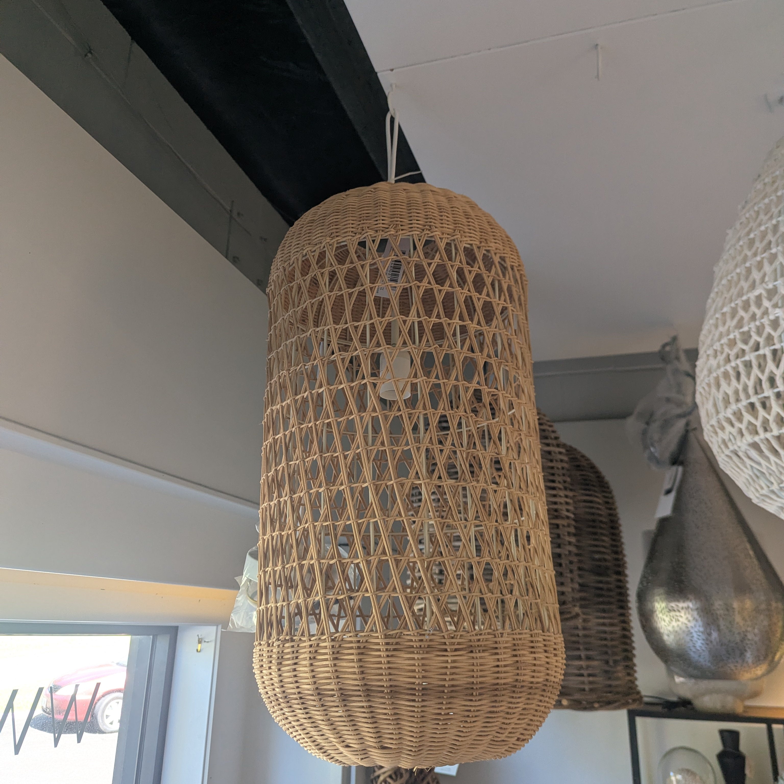 A large cylindrical woven rattan pendant light hanging in a modern room with a white ceiling and dark wooden beams.