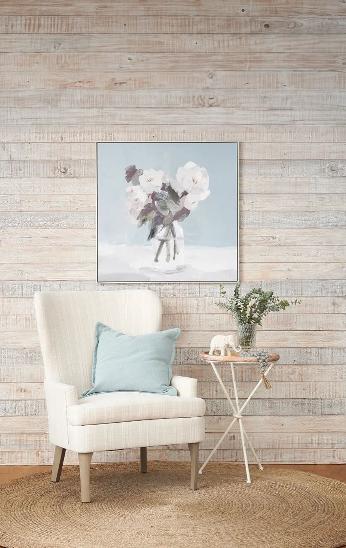 White armchair with a blue pillow in front of a wooden wall with a floral painting.