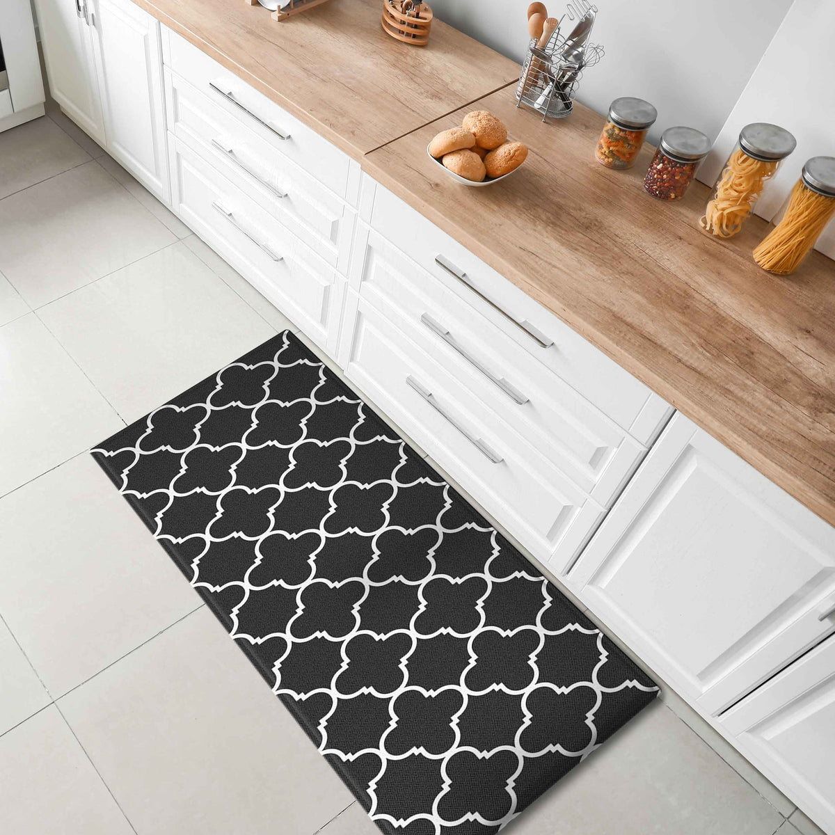 A black rectangular kitchen mat with a white geometric trellis pattern lies on the floor in front of white cabinets with light wood countertops. The scene is styled like a modern kitchen with jars of pasta and bread on the counter.