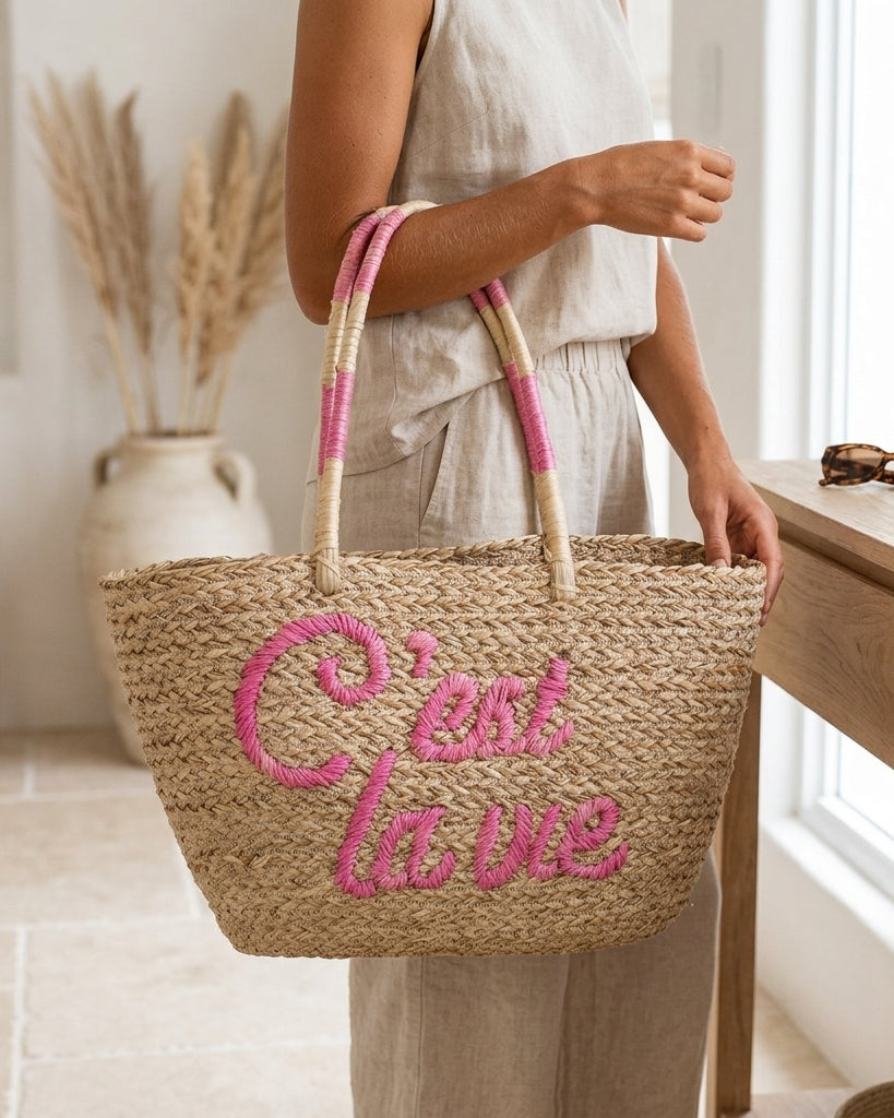 A woman in a light linen dress holds a natural woven straw tote bag with pink embroidered cursive text reading 'C'est la vie' and pink-and-beige striped handles, standing in a bright, neutral-toned room next to a wooden table.