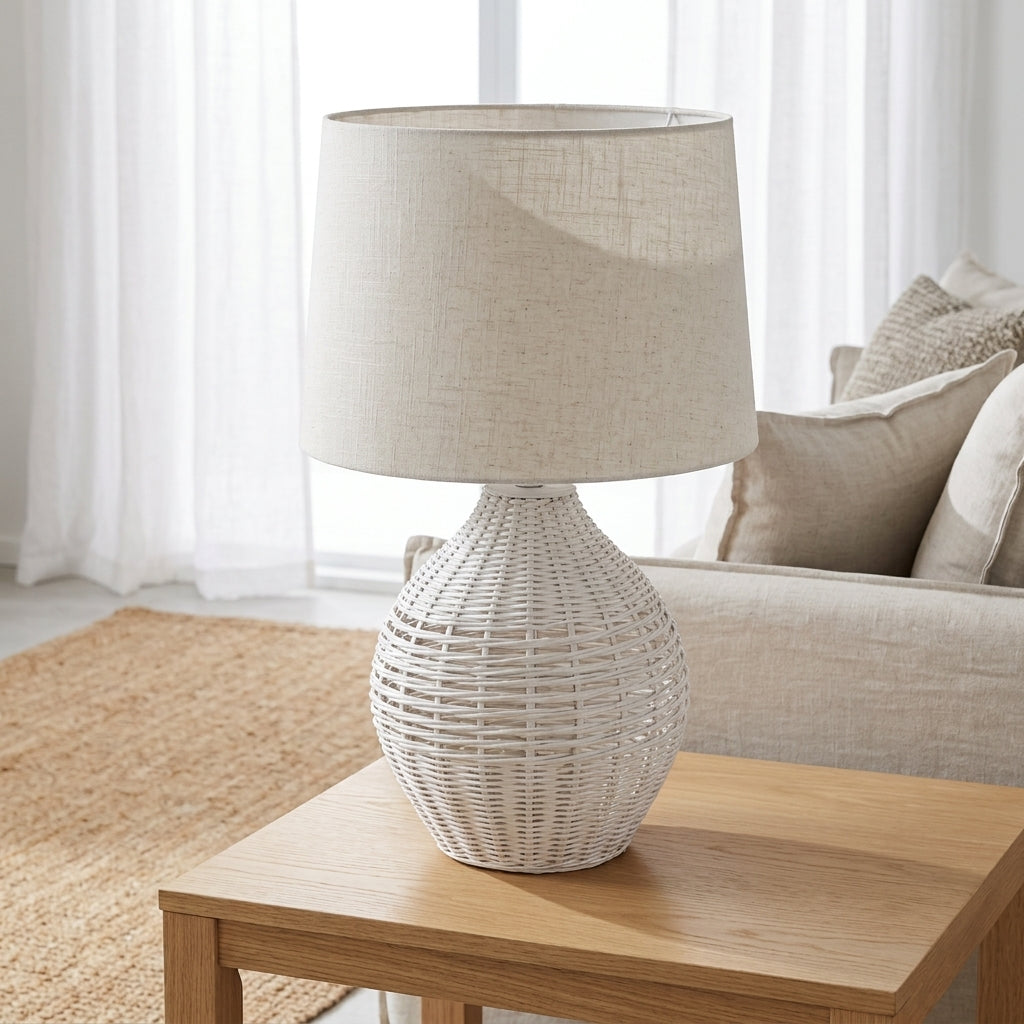 A white woven rattan table lamp with a beige linen drum shade sits on a light wood side table in a bright, modern living room setting.