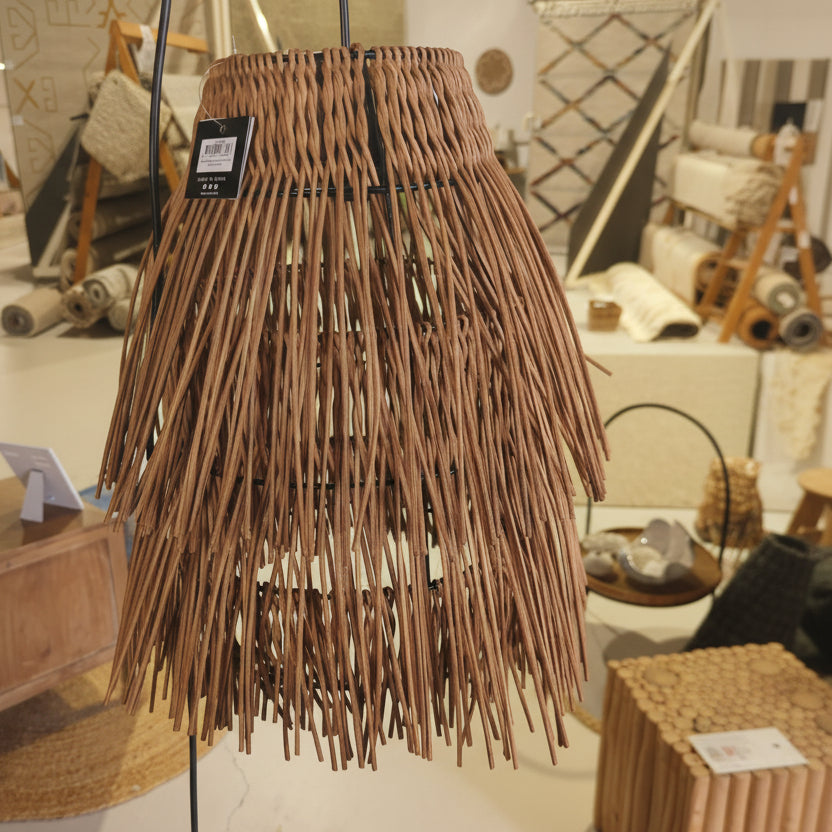 A woven wicker pendant light shade with long, thin brown strands hanging in a tapered conical shape, shown hanging indoors with a small product tag attached.