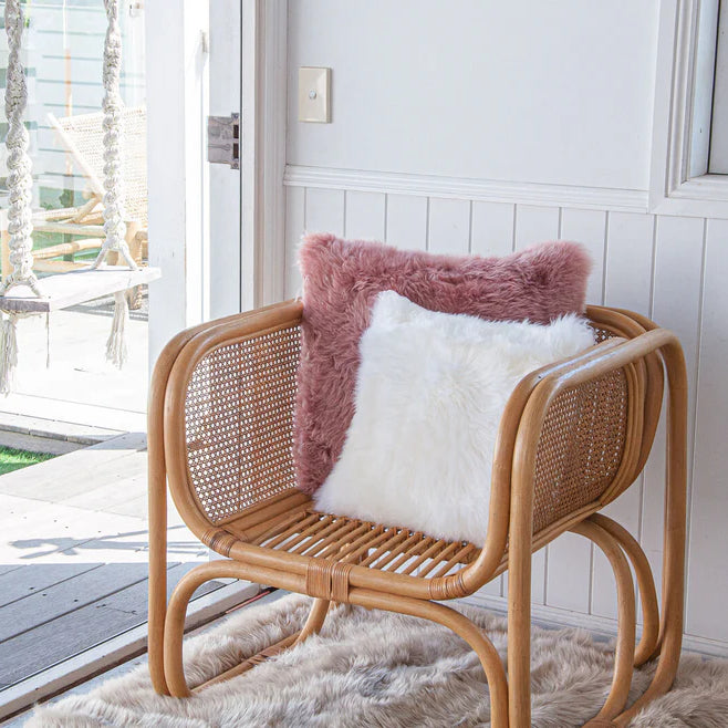 Rattan chair with pink and white sheepskin cushions on a patio with a view of a garden.