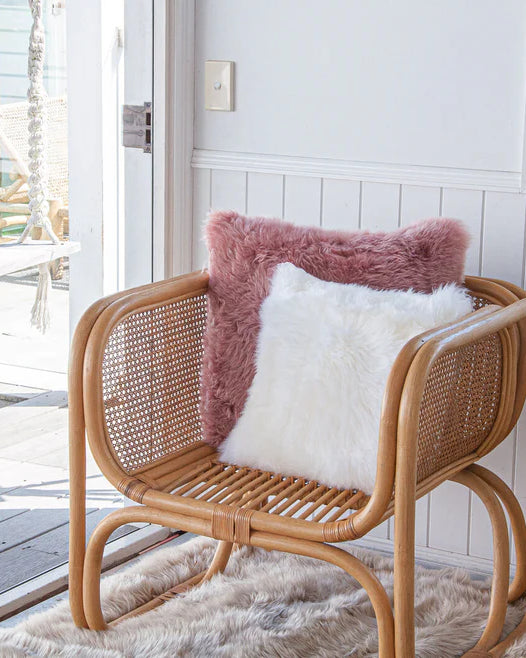 Rattan chair with pink and white sheepskin cushions on a patio with a view of a garden.