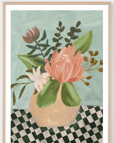 A framed art print featuring a still life painting of a pink protea flower in a beige vase, surrounded by green leaves, set against a light blue-green background with a black and white checkered surface.