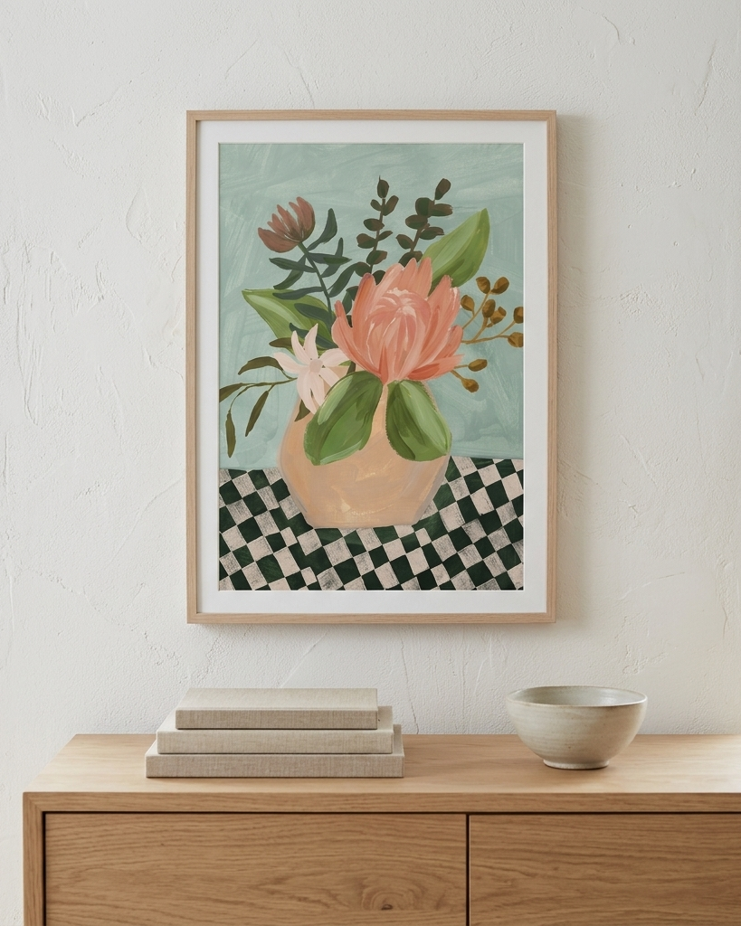 A framed floral art print featuring a pink flower in a beige vase against a light blue background, hung on a white textured wall above a wooden console table with books and a bowl.